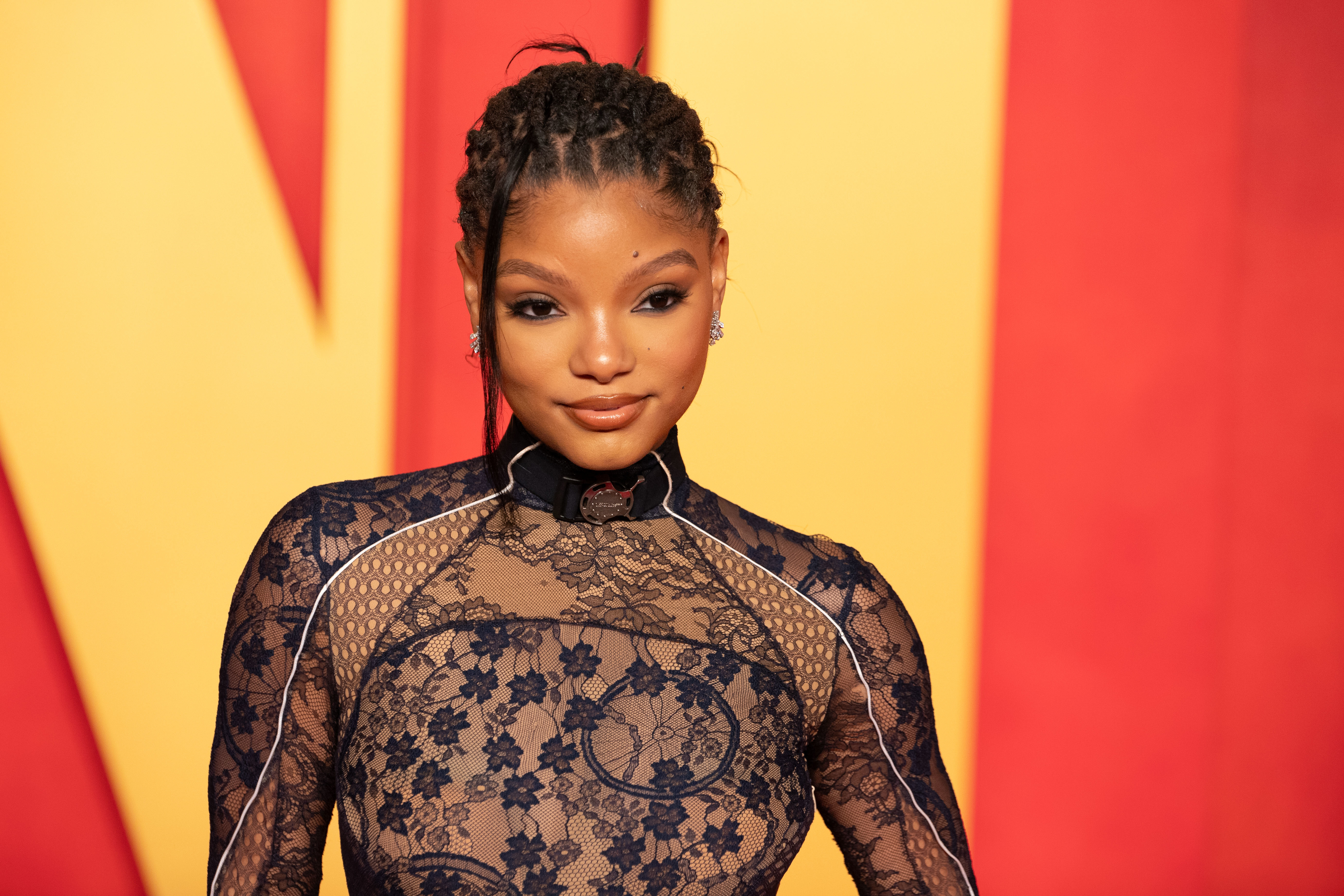 Halle Bailey on the red carpet, wearing a high-neck, sheer lace dress with intricate patterns