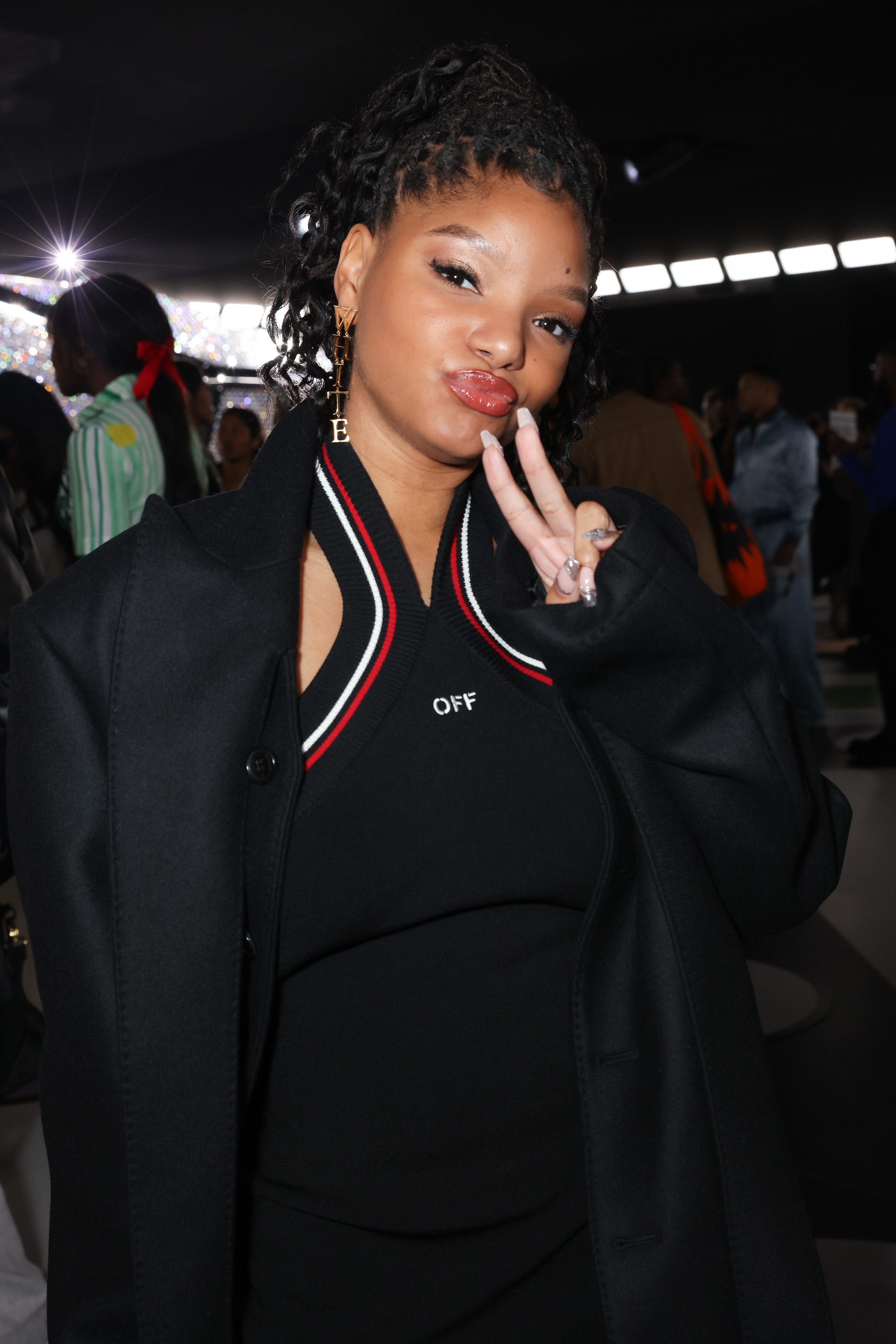 Halle Bailey at an event, wearing a black coat over a black top with white and red trim, making a peace sign with her fingers