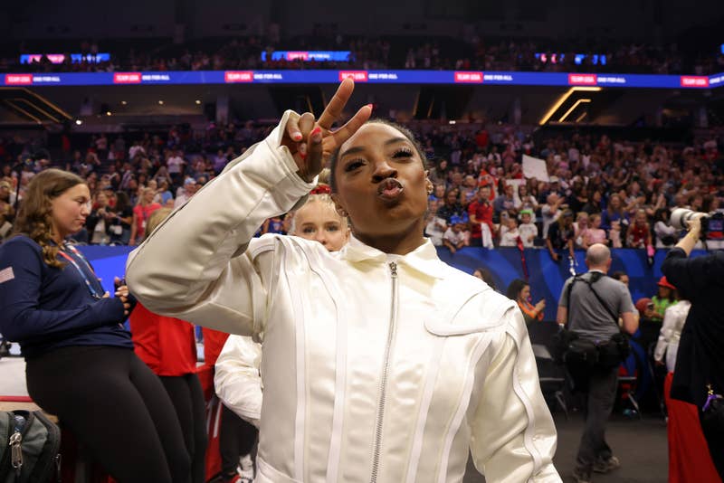 Simone Biles makes a peace sign and puckers her lips in front of a large crowd at an indoor event