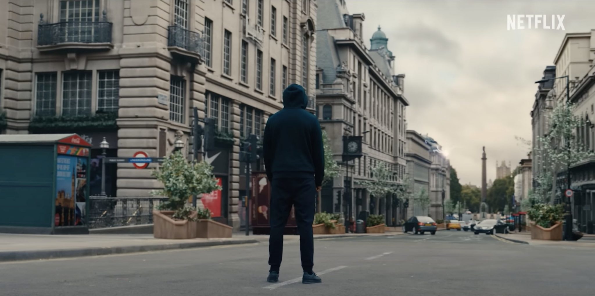 A person in a hoodie stands in the middle of an empty city street with historical buildings, seen in a scene from a Netflix show