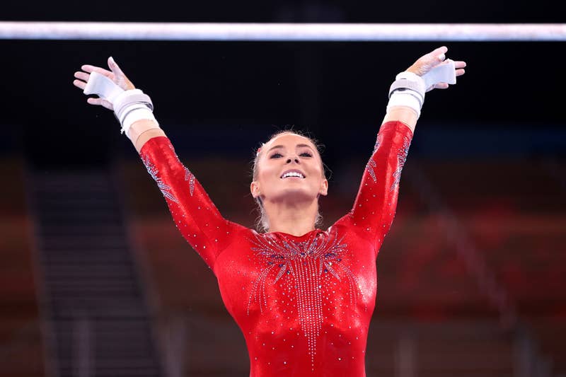 A gymnast raises her arms after a routine in an empty arena, wearing a sparkly leotard with a pattern on the front