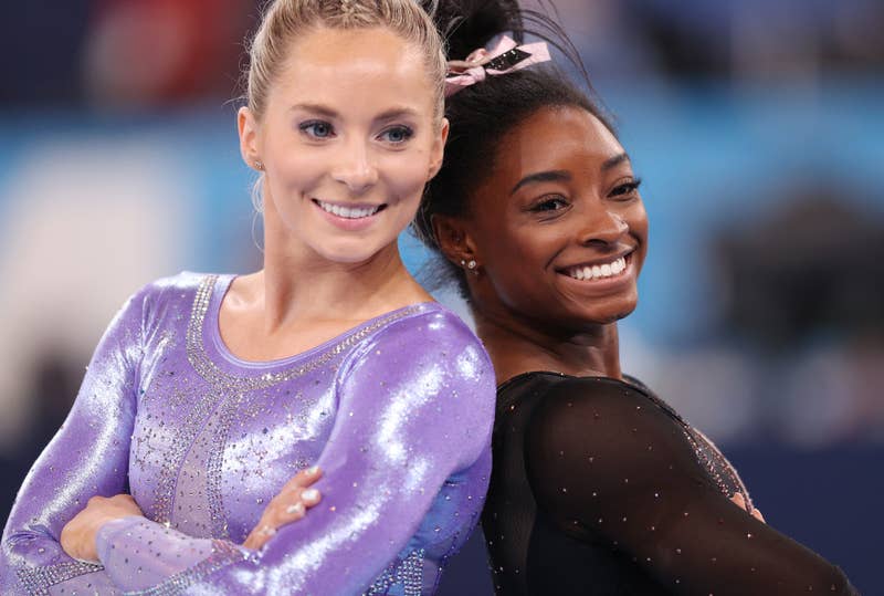 Simone Biles and another gymnast stand back-to-back, each smiling and wearing sparkly leotards