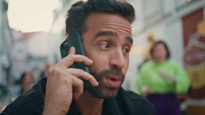 Man talking on a phone pressed to his ear in an urban setting; a blurred figure in the background
