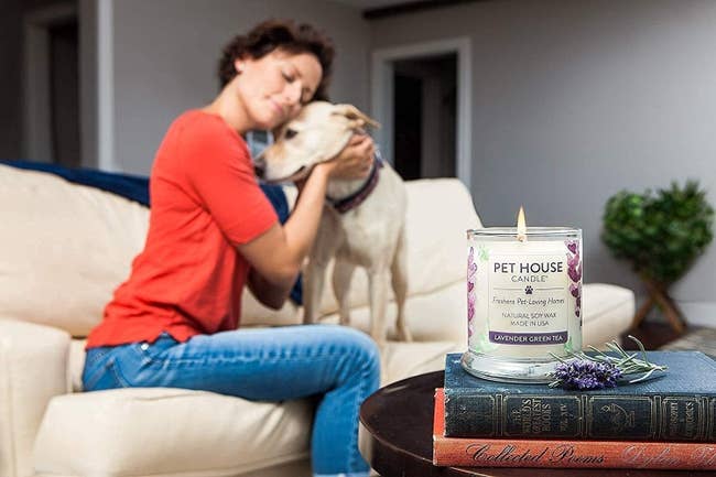 A person embraces a dog on a couch. In the foreground, a Pet House candle labeled
