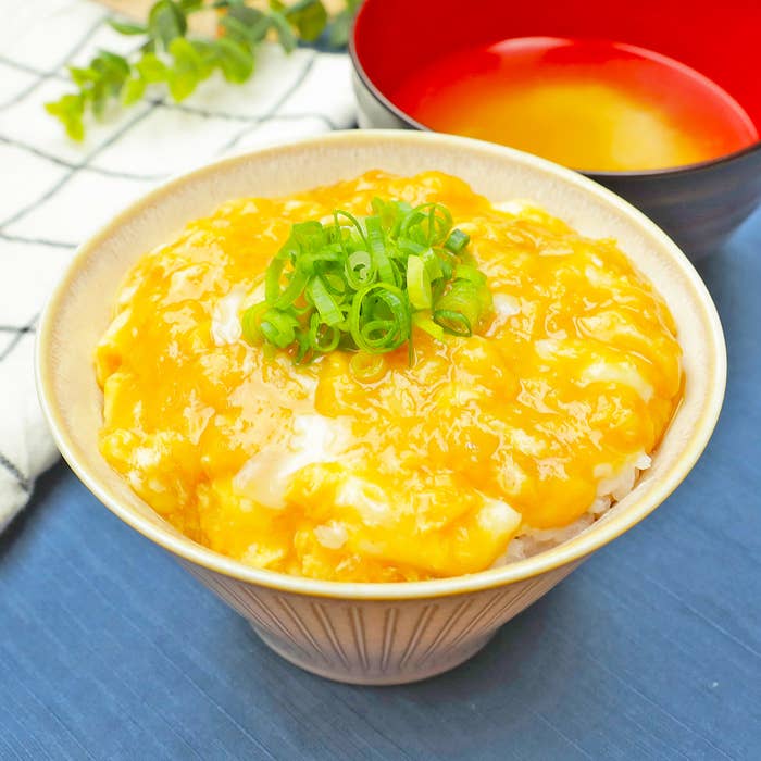 丼に卵とじを載せたご飯、ネギがトッピングされている。後ろに味噌汁が並んでいる。