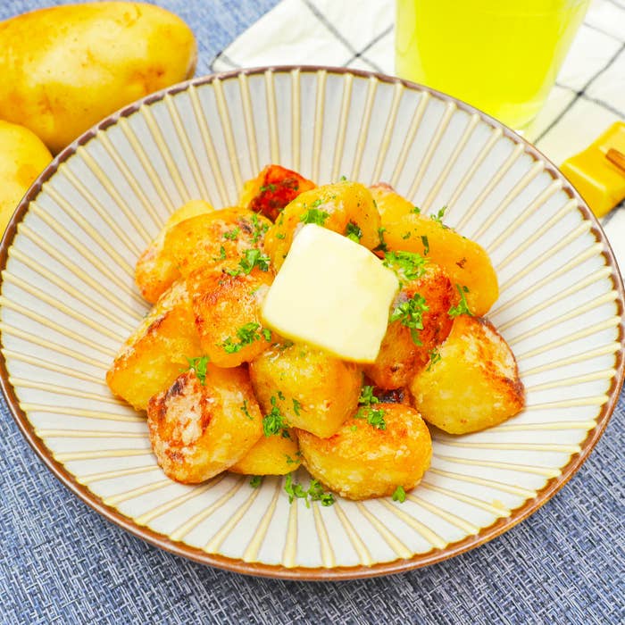 お皿に盛られた焼きポテトの上にバターと刻んだパセリが散らされています。背景にはじゃがいもと黄色の飲み物があります。