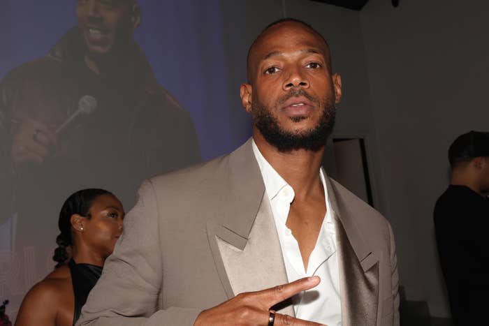 Marlon Wayans at an event wearing a formal suit, holding up a peace sign, with a projected image of himself in the background
