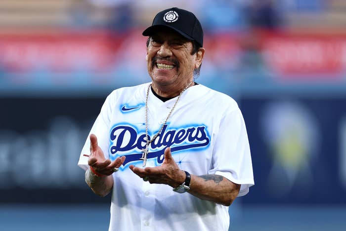 Danny Trejo smiles widely while wearing a Los Angeles Dodgers jersey and a cap at a Dodgers game