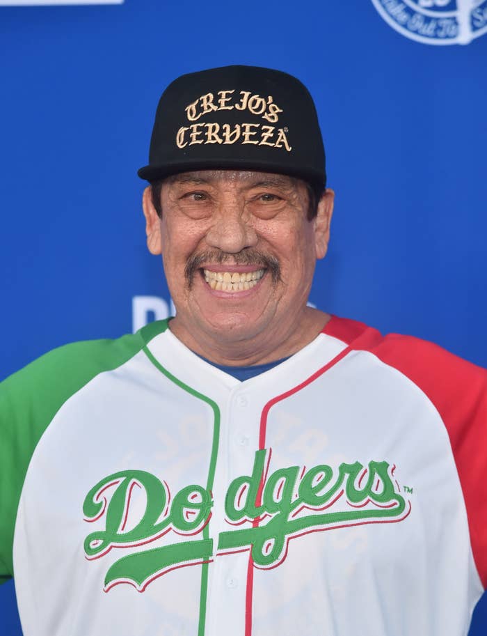 Danny Trejo is smiling, wearing a baseball cap that reads "Trejo's Cerveza" and a baseball jersey with "Dodgers" on it