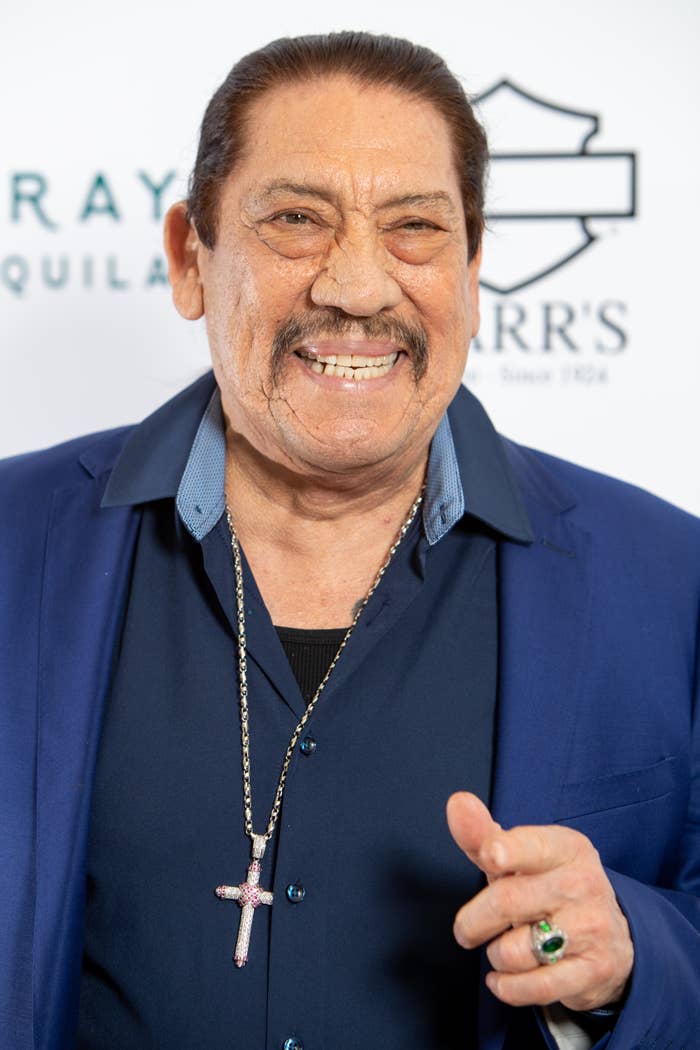 Danny Trejo wearing a blue jacket and a cross necklace, smiling at a step and repeat event