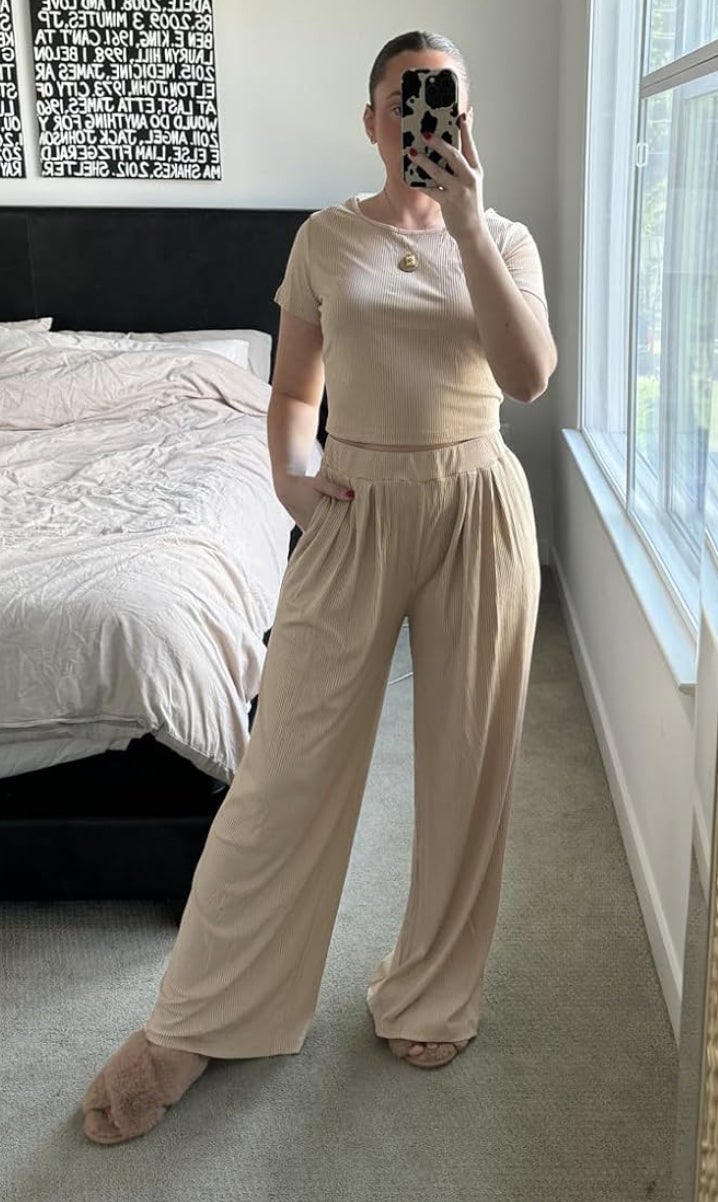 Person in a mirror selfie wearing a casual loungewear set with a T-shirt and wide-legged pants, one hand in pocket, one foot wearing a slipper. Background: unmade bed