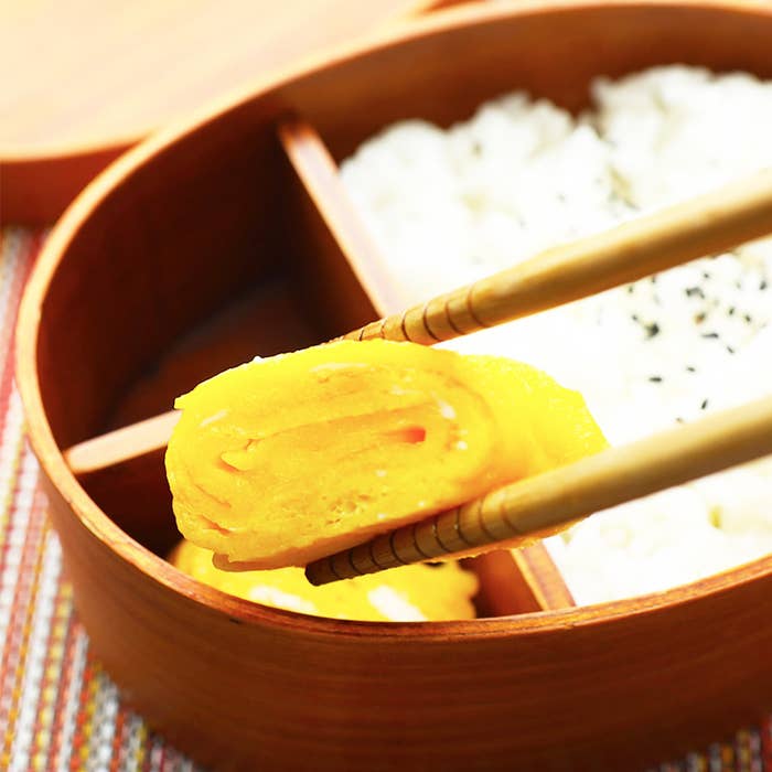 箸で持ち上げられた卵焼きとご飯が詰められた木製の弁当箱。