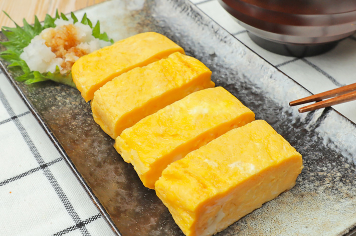 長方形の皿に盛られた4つの卵焼きと、おろししょうが、大葉の盛り合わせ。横にお椀と箸も見える。