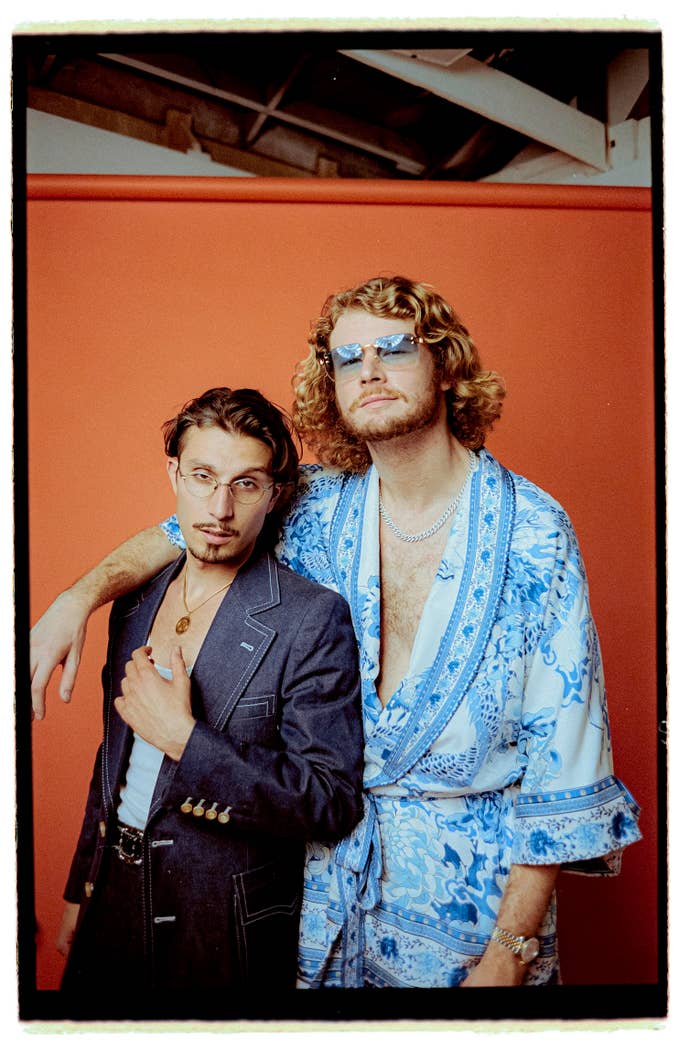 Two men pose together, one wearing a suit with an open collar and glasses, the other in a robe with intricate patterns and sunglasses. Names unknown