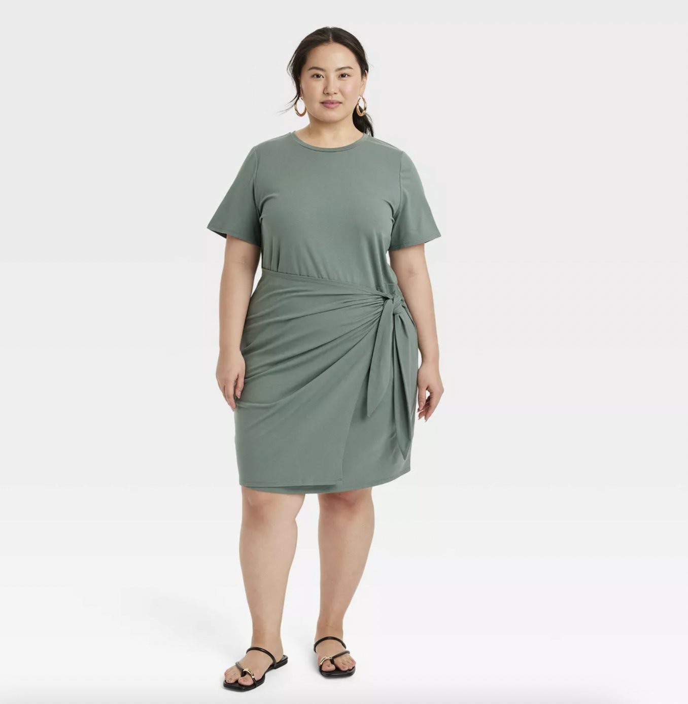 A person wearing a short-sleeved dress with a knot detail on the side and sandals stands against a plain background