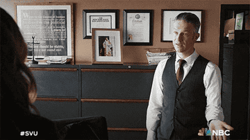 A man in a vest and tie gestures while talking to a woman in an office with framed certificates on the wall. Two acronyms, &quot;SVU&quot; and &quot;NBC,&quot; are in the corners