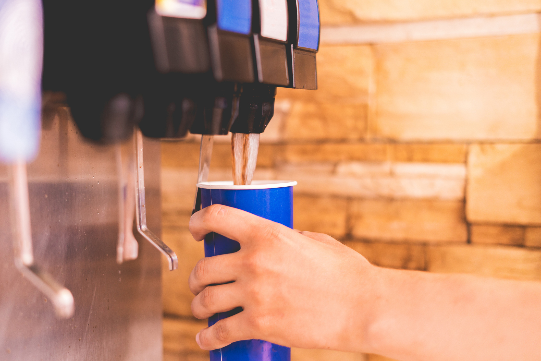 A hand is seen filling a blue cup with a drink from a soda fountain dispenser against a tiled background