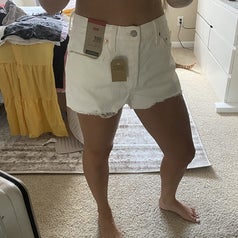 A person showcases white denim shorts with tags still attached. The person is in a bedroom with assorted clothes on the bed and floor