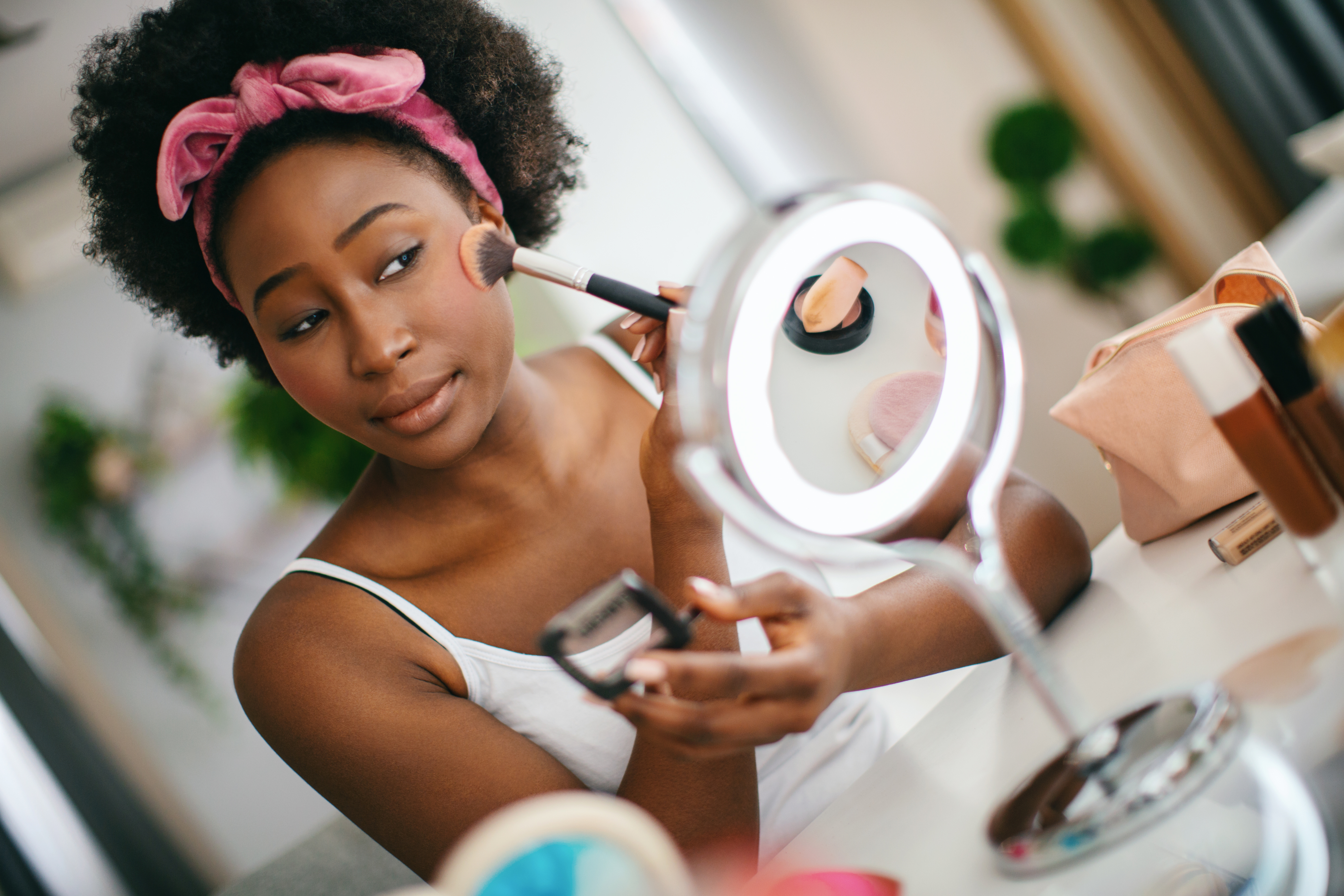 Woman applies makeup with a brush while looking in a mirror at her vanity table. She wears a white camisole and a pink headband