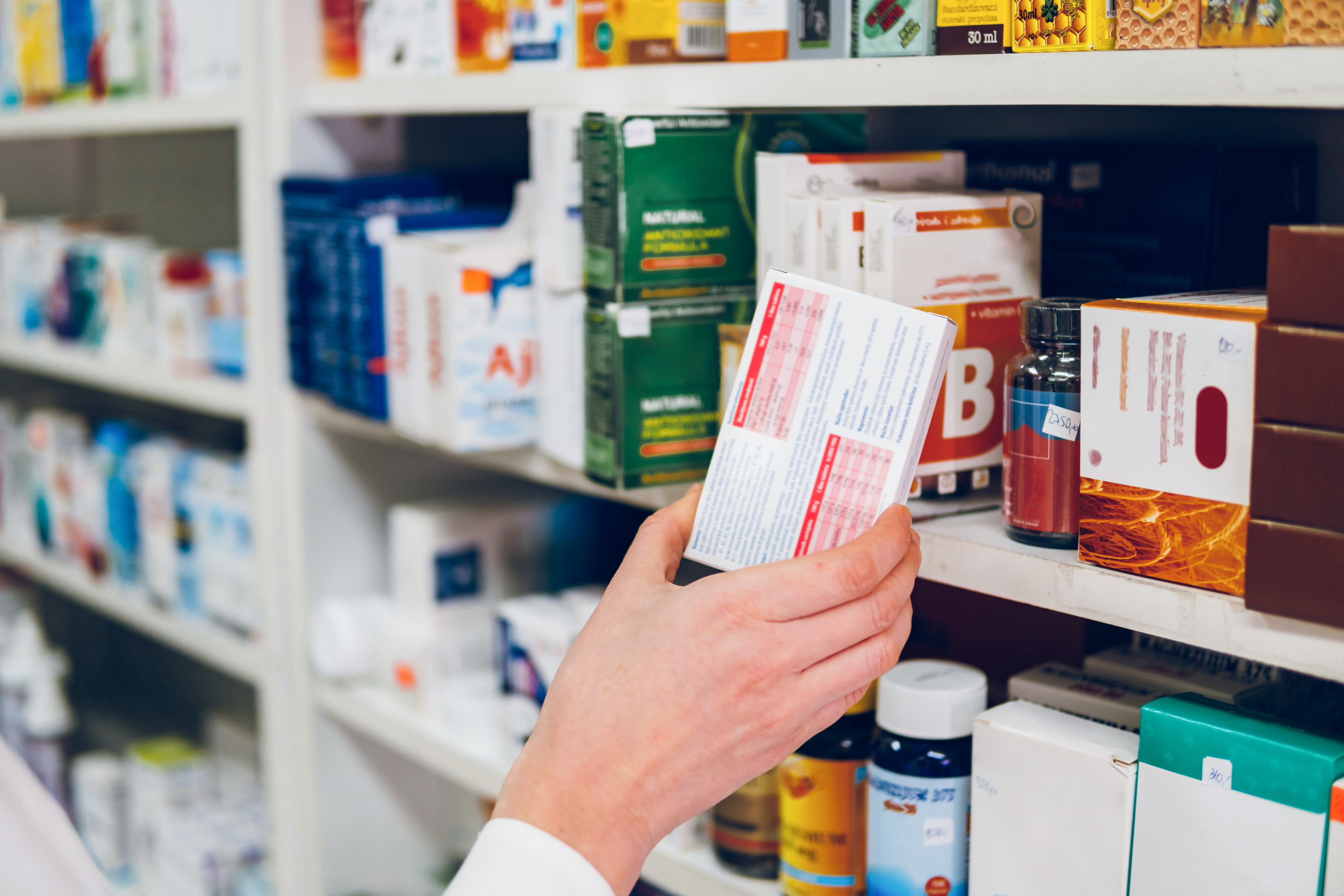 Close-up of a person's hand holding a boxed medication in a pharmacy. Shelves in the background are stocked with various health products and medications