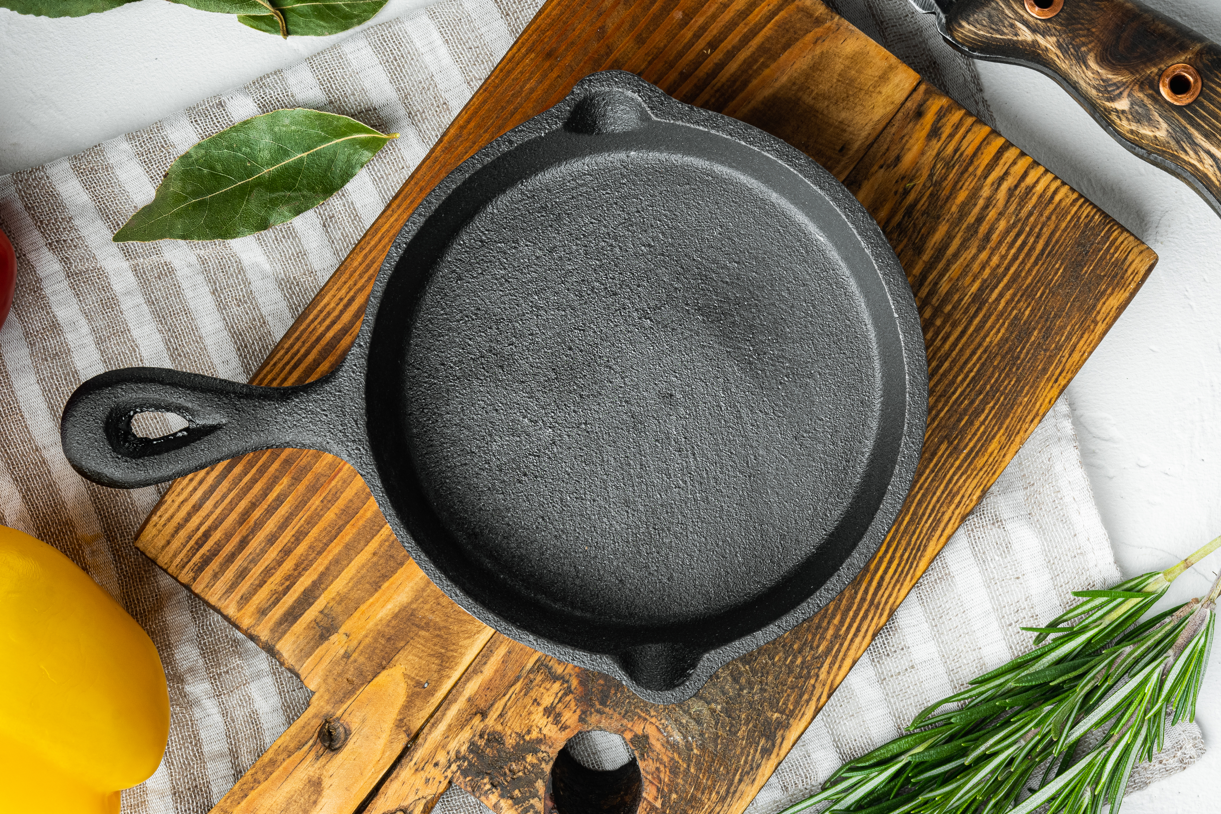 A black cast-iron skillet sits on a wooden board and a striped cloth, surrounded by bell peppers, a knife, and fresh herbs like rosemary and bay leaves