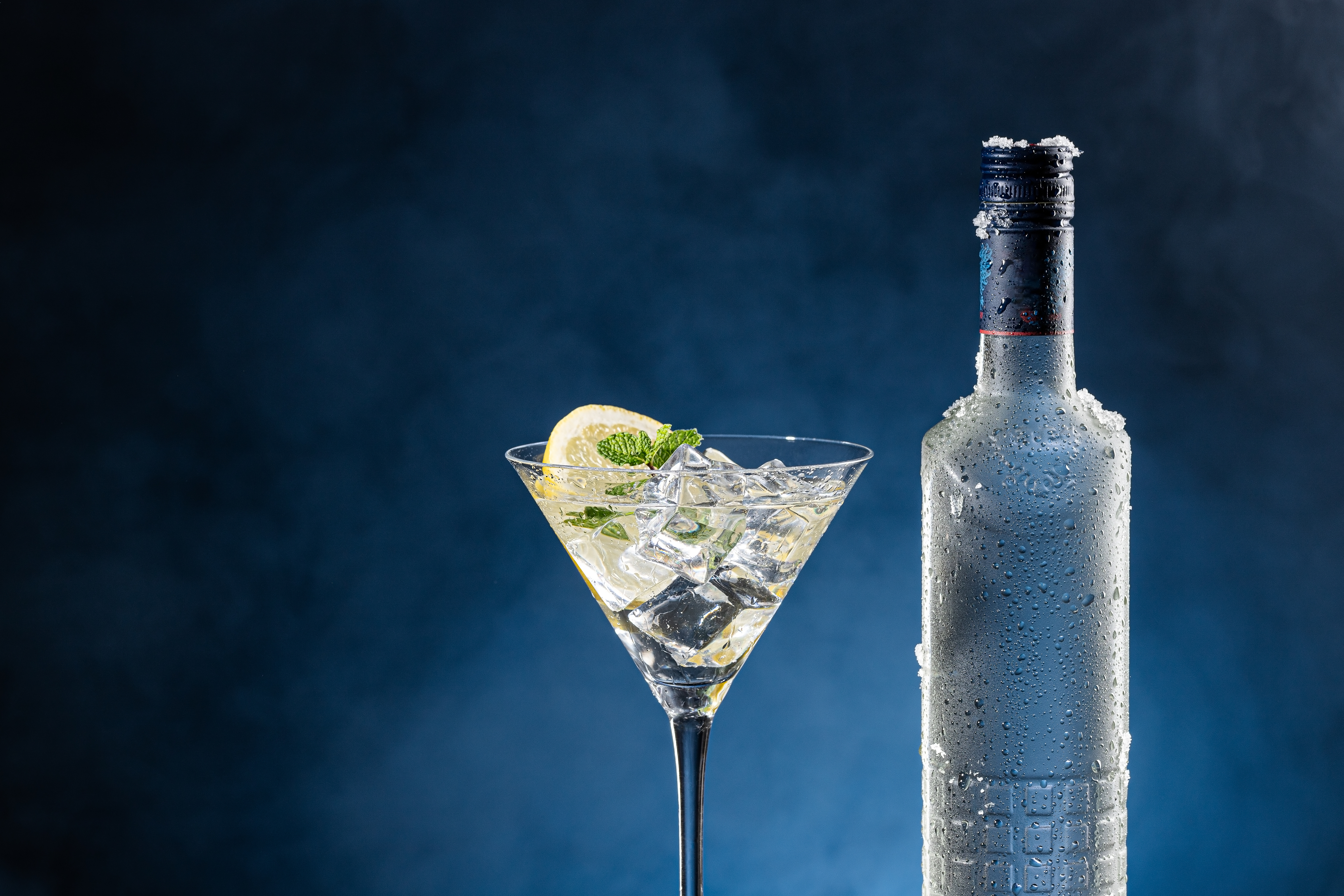 A frosty vodka bottle stands next to a martini glass filled with ice, a lemon wedge, and a mint garnish in a work and money-themed image