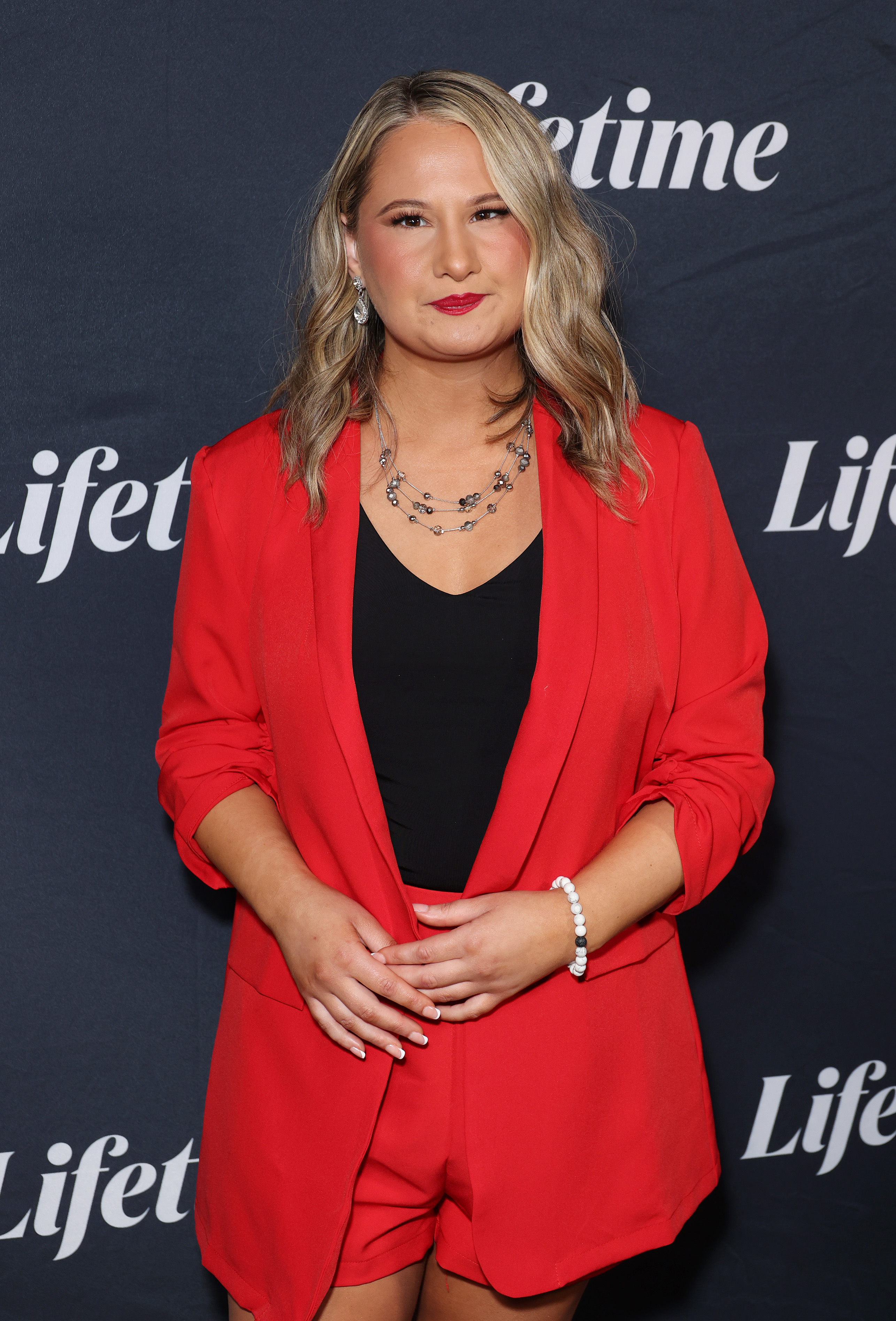 A woman in a red suit jacket and shorts stands in front of a Lifetime backdrop, smiling