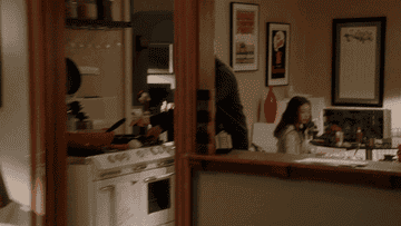 gif of A man cooks on a stove while a girl sits at a nearby table in a kitchen. Movie posters and decor are visible in the background
