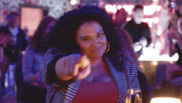 gif A person with curly hair is smiling and pointing at the camera in a busy, lively indoor setting with people in the background