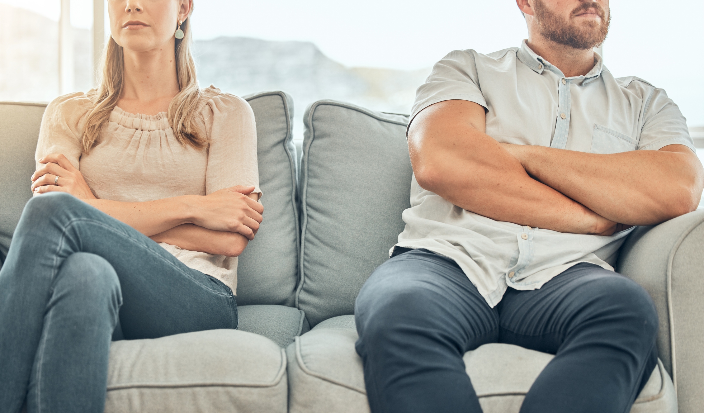 A woman and a man sit on a couch with arms crossed, looking away from each other, appearing to be in conflict