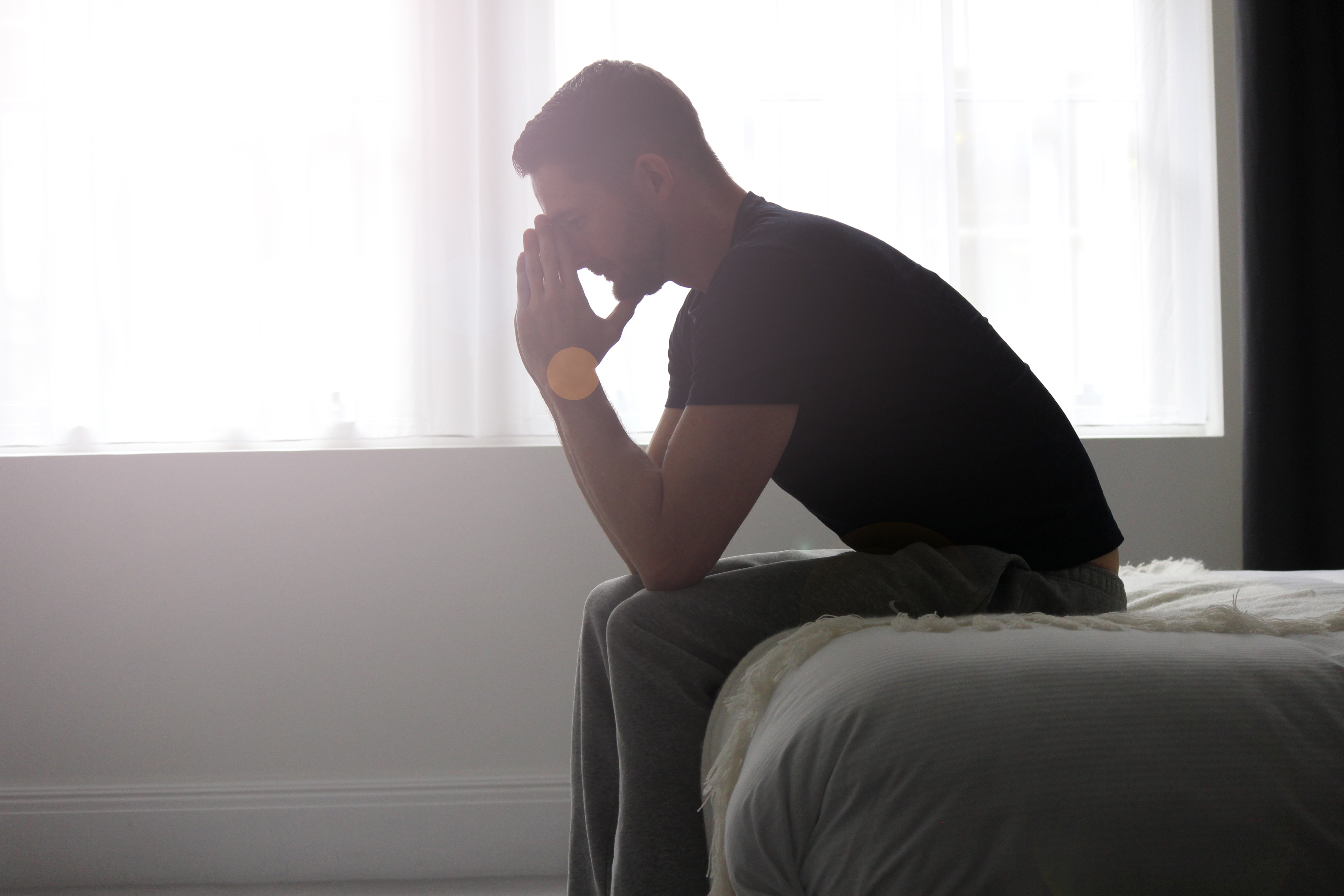 A man sits on the edge of a bed in a pensive posture, with hands clasped near his face, in a softly lit room