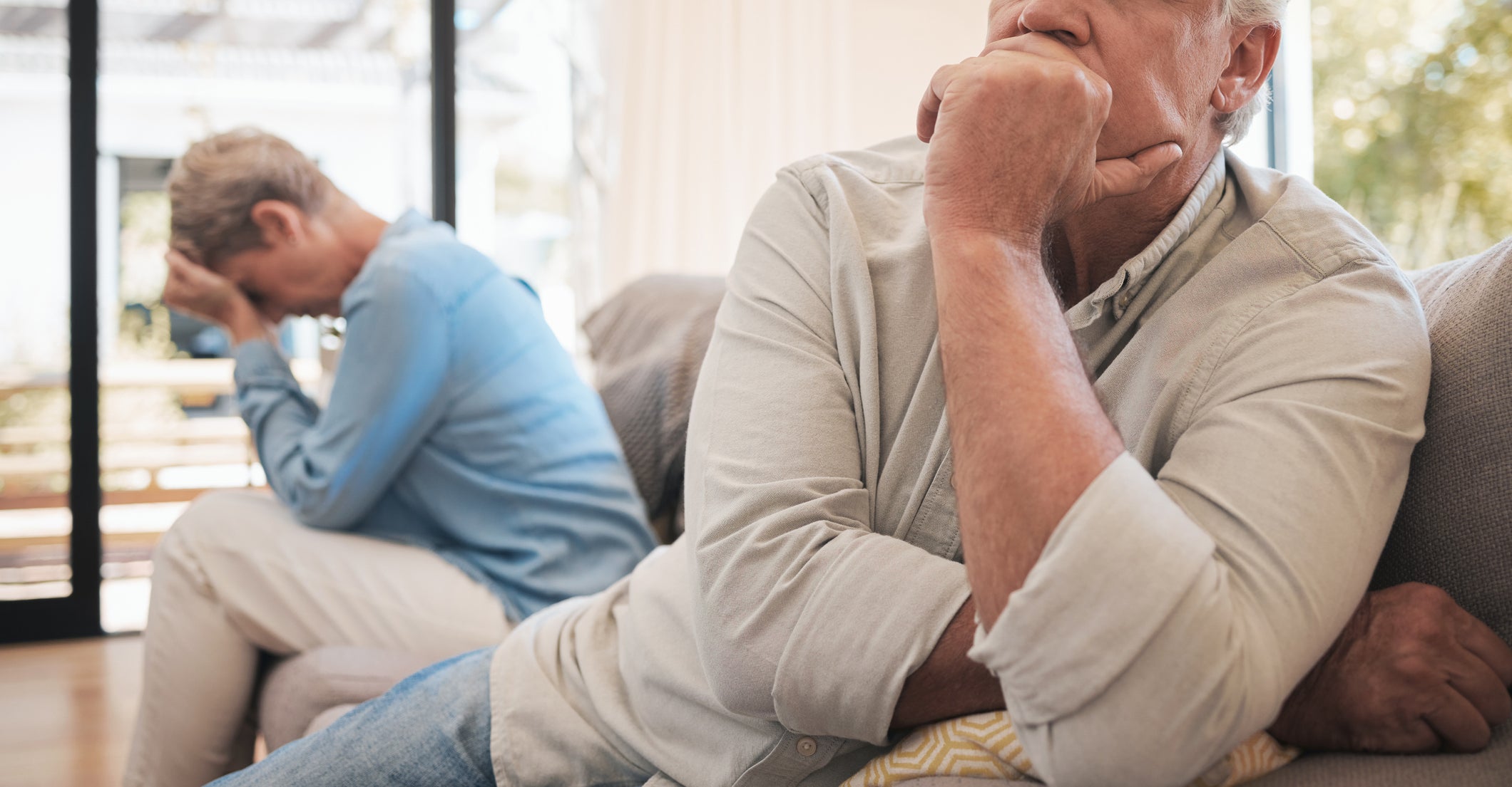 An older man and woman sit on a couch, facing away from each other, both looking distressed. The man is in the foreground, while the woman covers her face