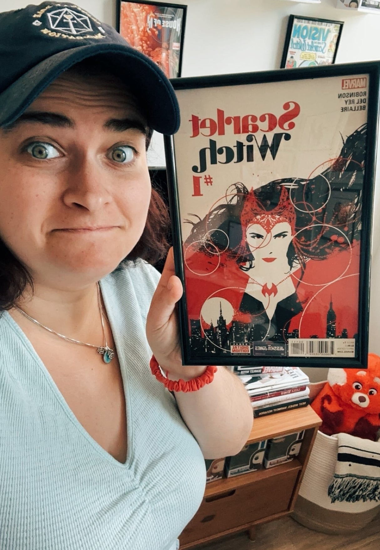 Nora, wearing a cap, holds up a framed Scarlet Witch comic book