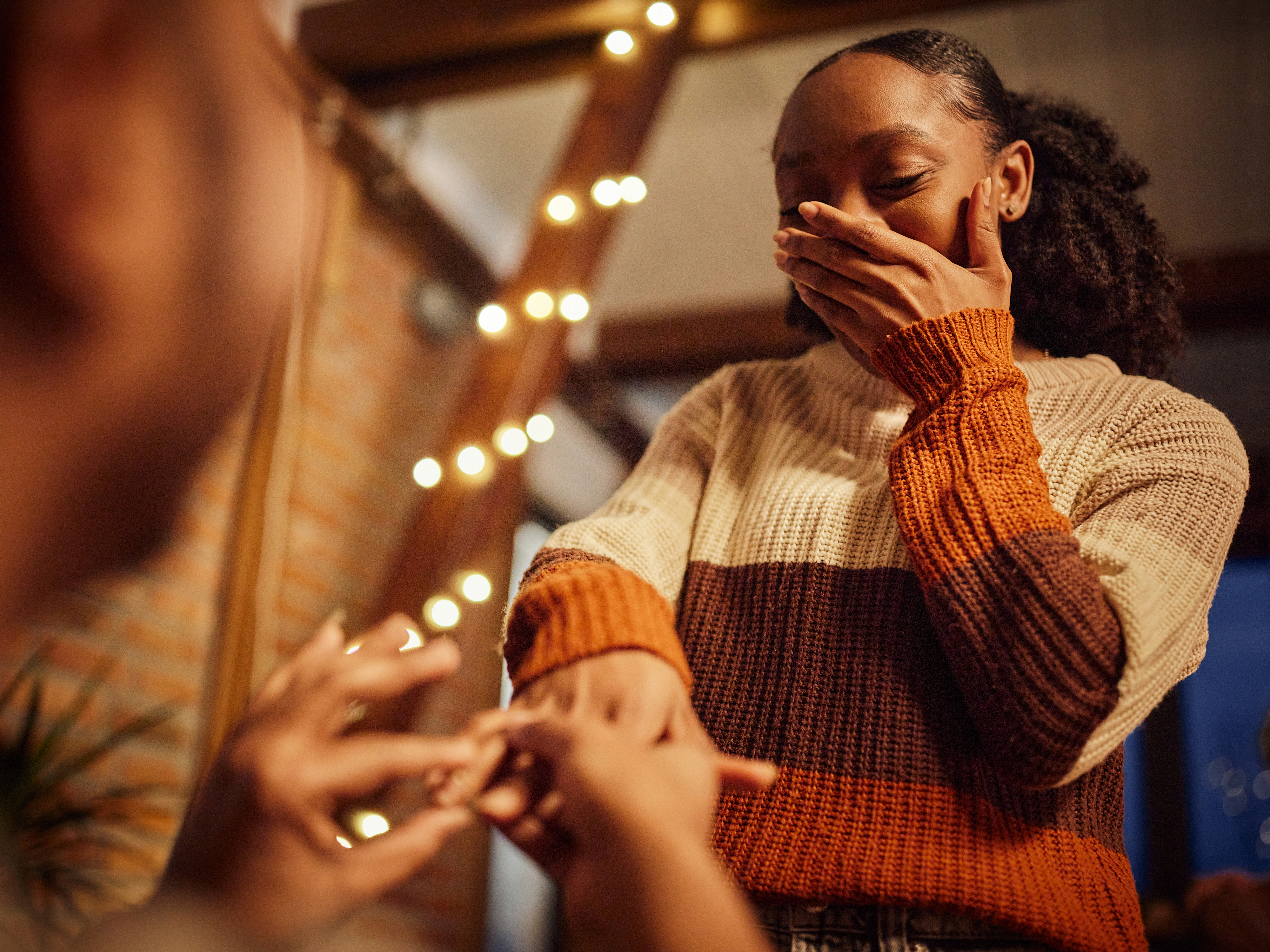 Person covering their mouth in surprise during a marriage proposal; they are indoors with string lights in the background