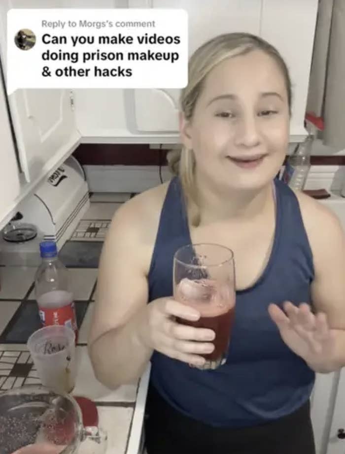 A woman in a kitchen holds a glass and smiles at the camera. A comment overlay asks, "Can you make videos doing prison makeup & other hacks?"