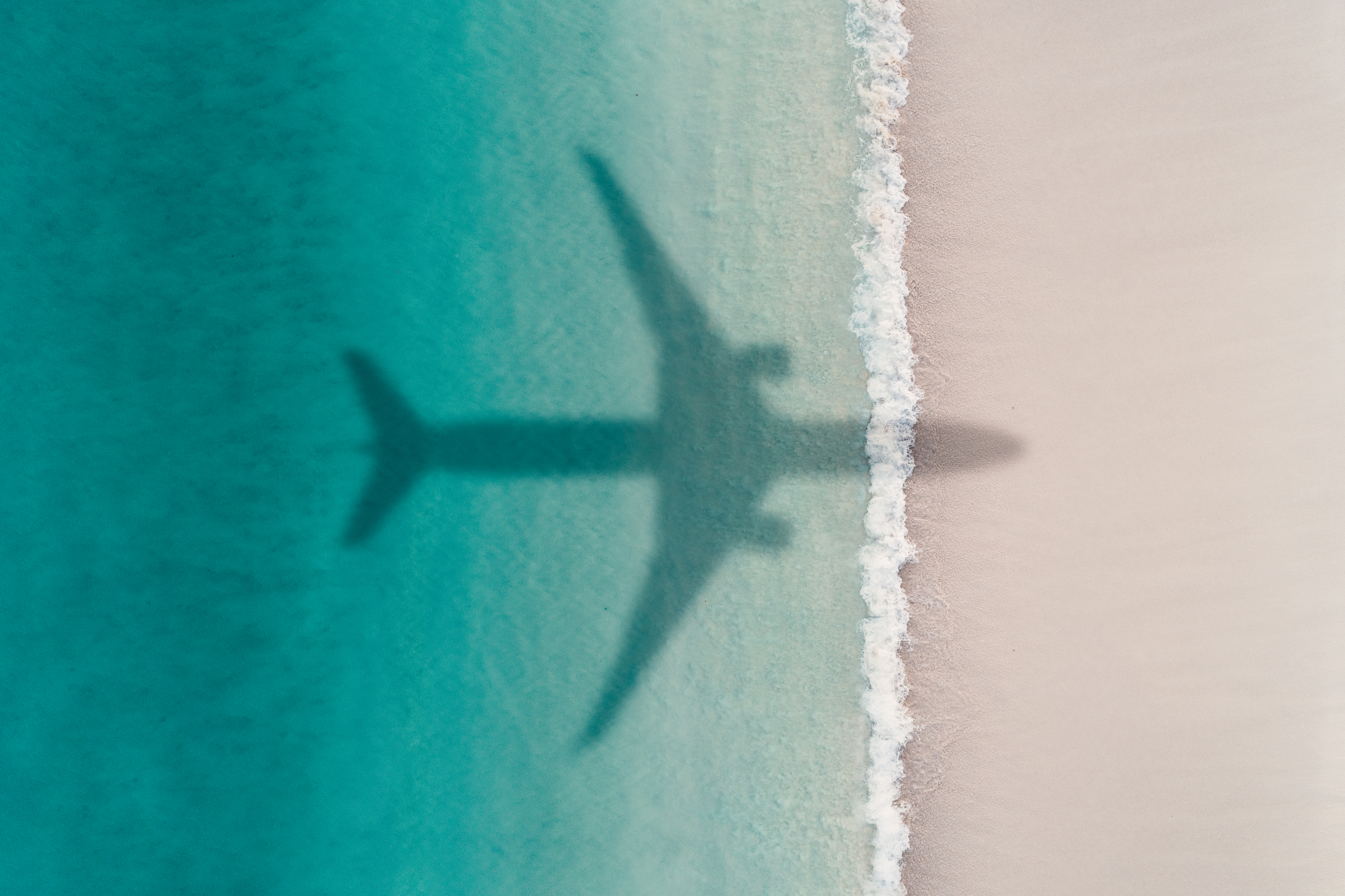 Aerial view of an aircraft's shadow over turquoise water and sandy shore, highlighting the interplay between travel and the natural landscape