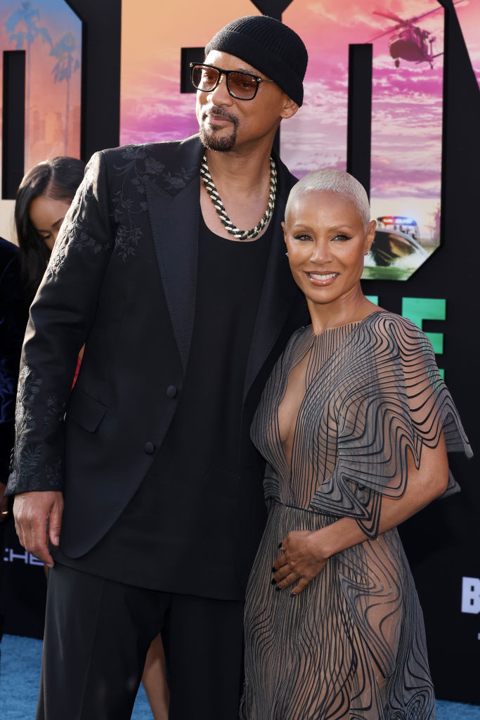 Will Smith and Jada Pinkett Smith at an event. Will is wearing a black hat, sunglasses, a black suit, and a chain necklace. Jada is in a sheer, wavy-patterned dress
