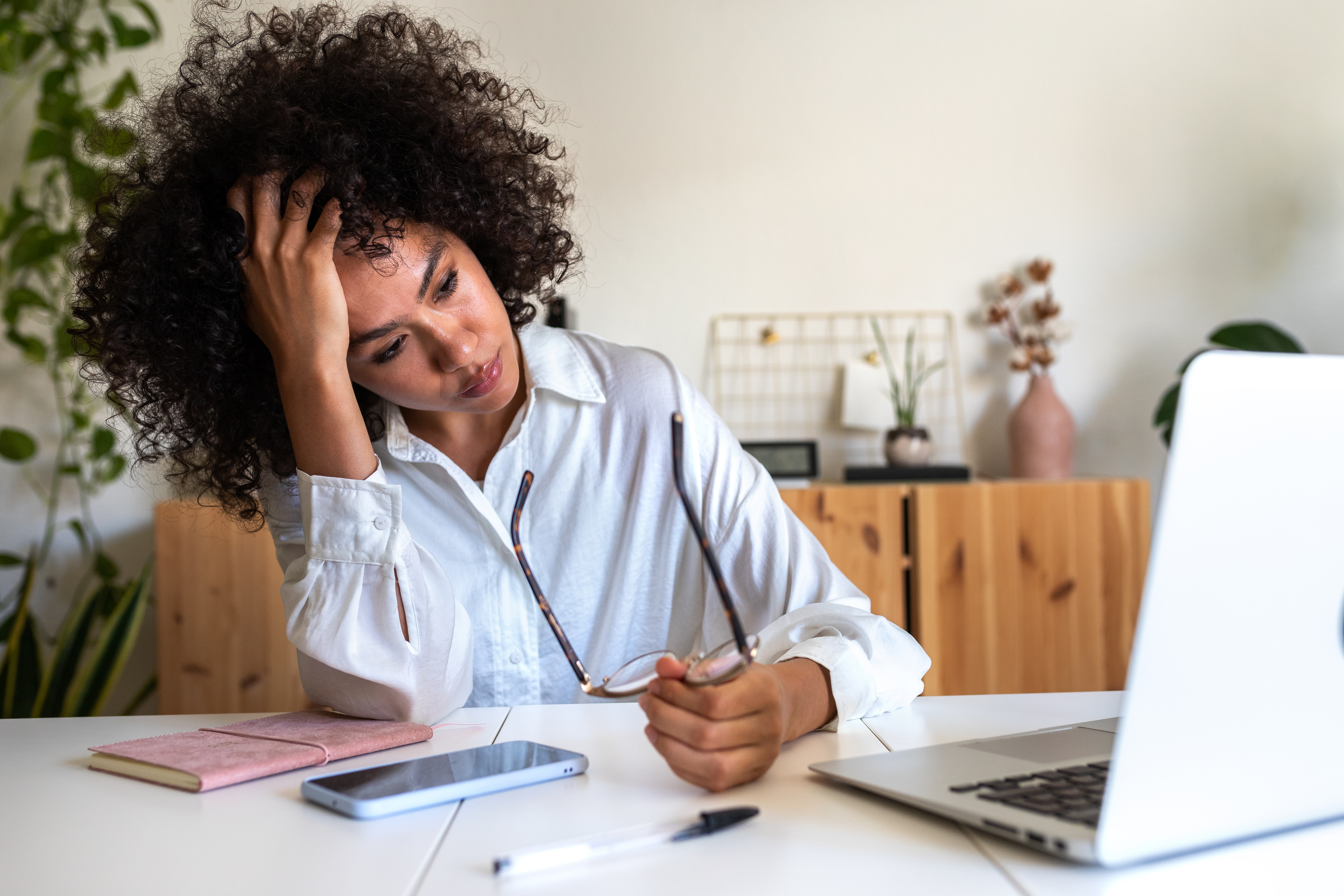 A woman with curly hair in a white blouse, seated at a desk with a laptop, notebook, phone, and a pen, looking stressed while holding her glasses