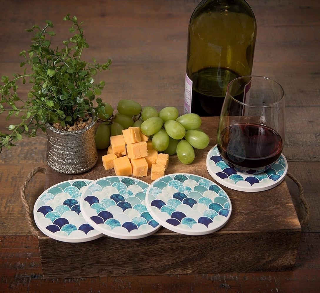 Coasters with a blue and white wave pattern on a wooden tray, a glass of red wine, a wine bottle, green grapes, cheese cubes, and a small plant