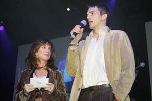 Kathy Griffin and Ashton Kutcher on stage. Kathy holds note cards and wears a velvet jacket. Ashton holds a microphone, dressed in a blazer and light shirt