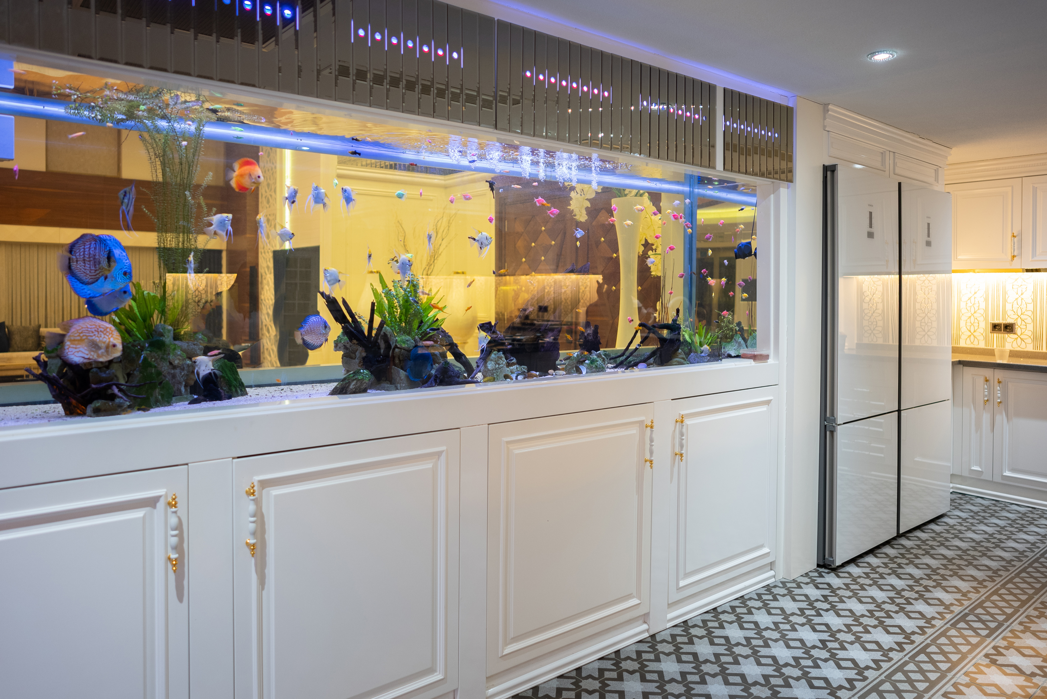 A spacious kitchen features an elegant, long fish tank with various colorful fish. The room includes white cabinets, a large refrigerator, and patterned flooring