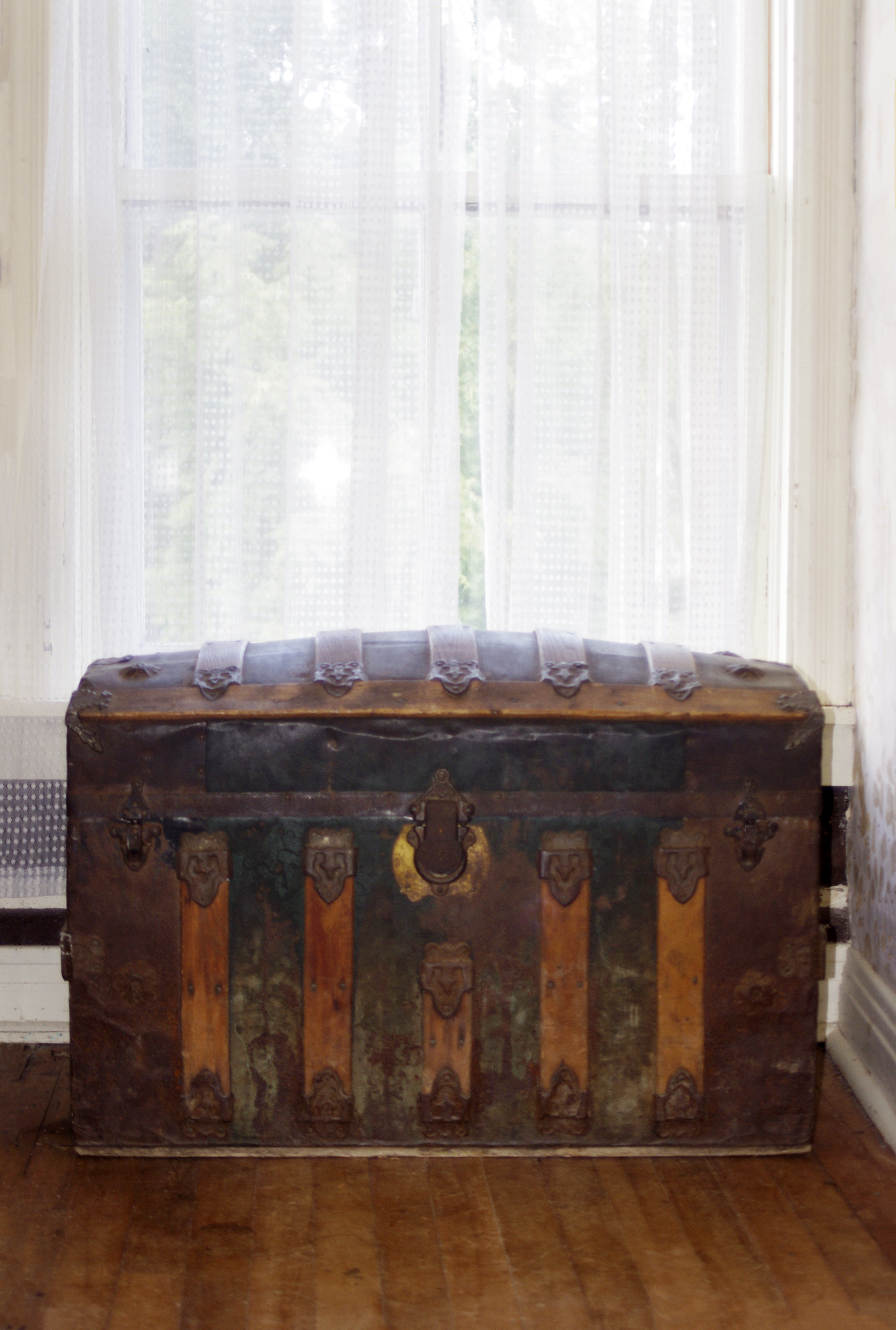 An old, intricately designed wooden trunk sits on a wooden floor in front of sheer curtains covering a window