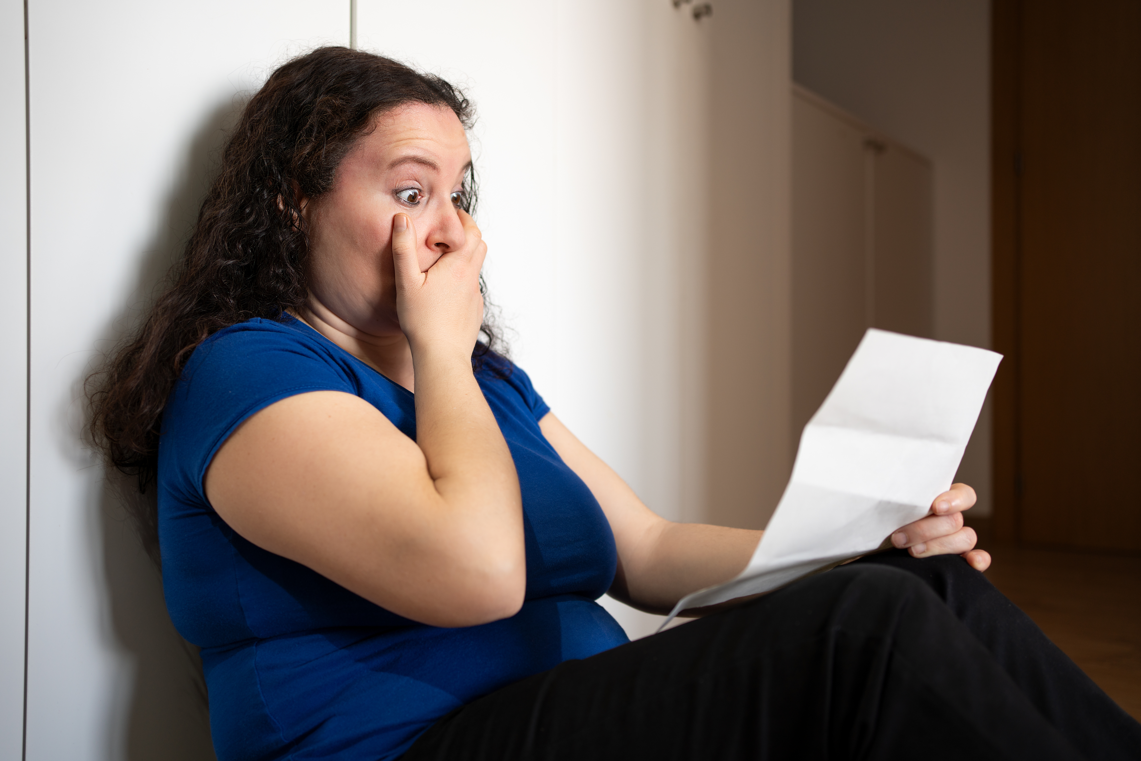 A woman in a blue t-shirt looks shocked while sitting against a wall, holding a piece of paper