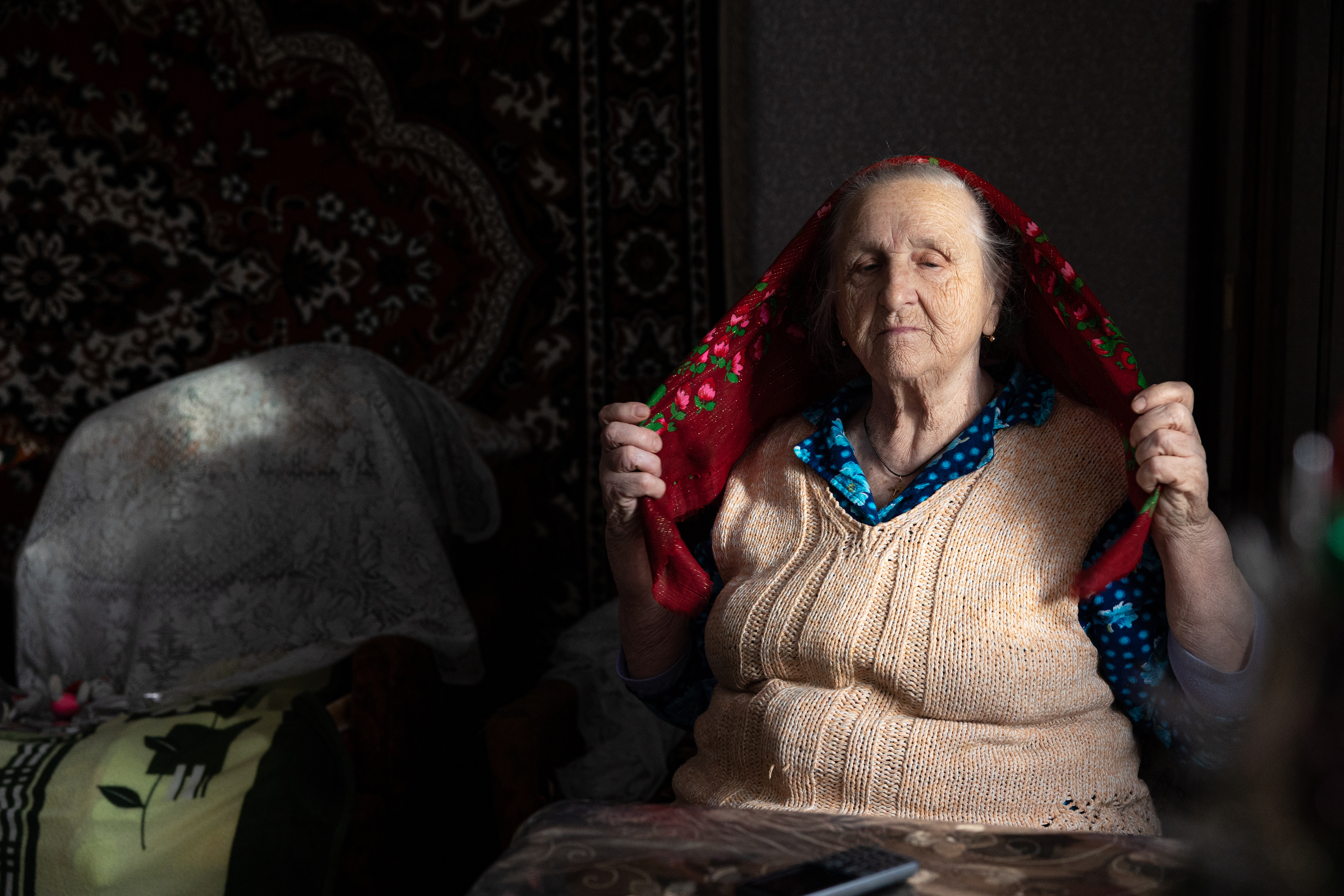 An elderly woman sits in a sunlit room, lifting a patterned scarf over her head with a peaceful expression