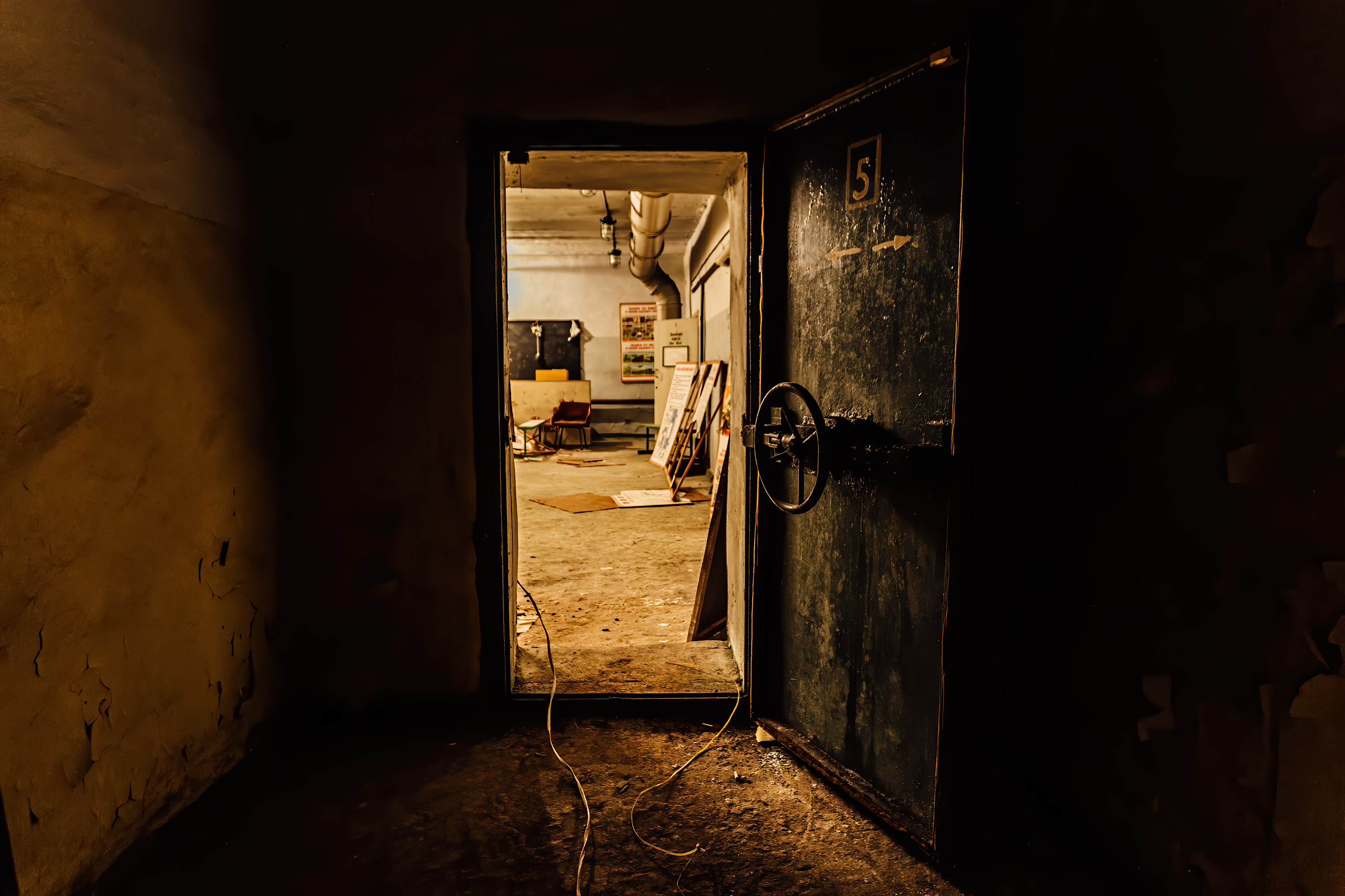 View of a dimly lit, industrial room with an open metal door. Various items and equipment are scattered in the background, suggesting possible disuse or storage