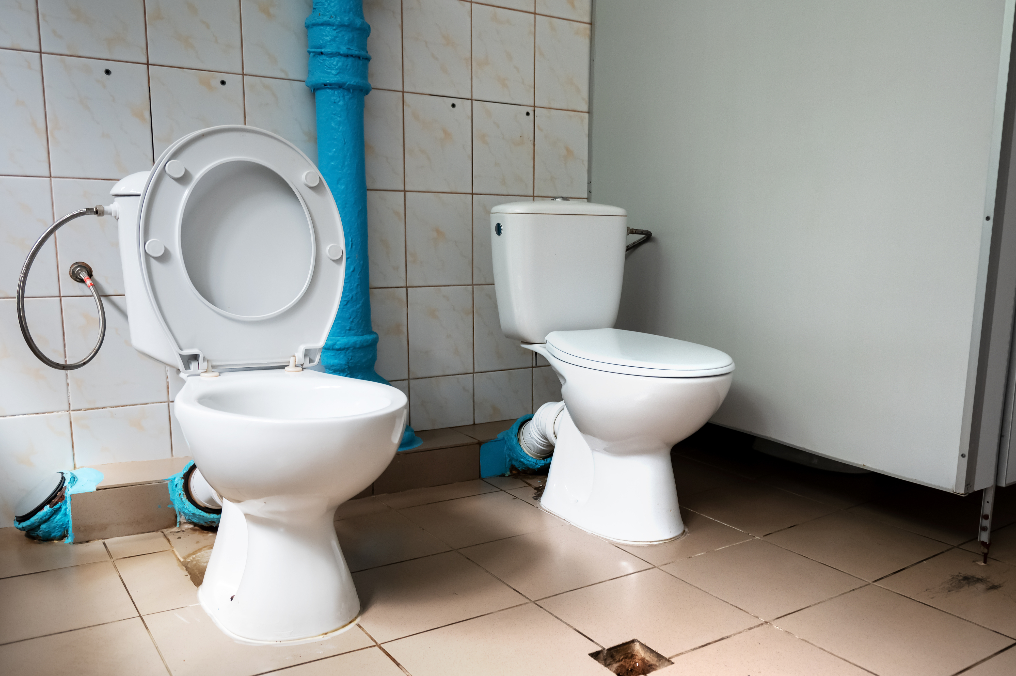 Two adjacent toilets in a shared restroom with tile walls and floors, featuring exposed plumbing