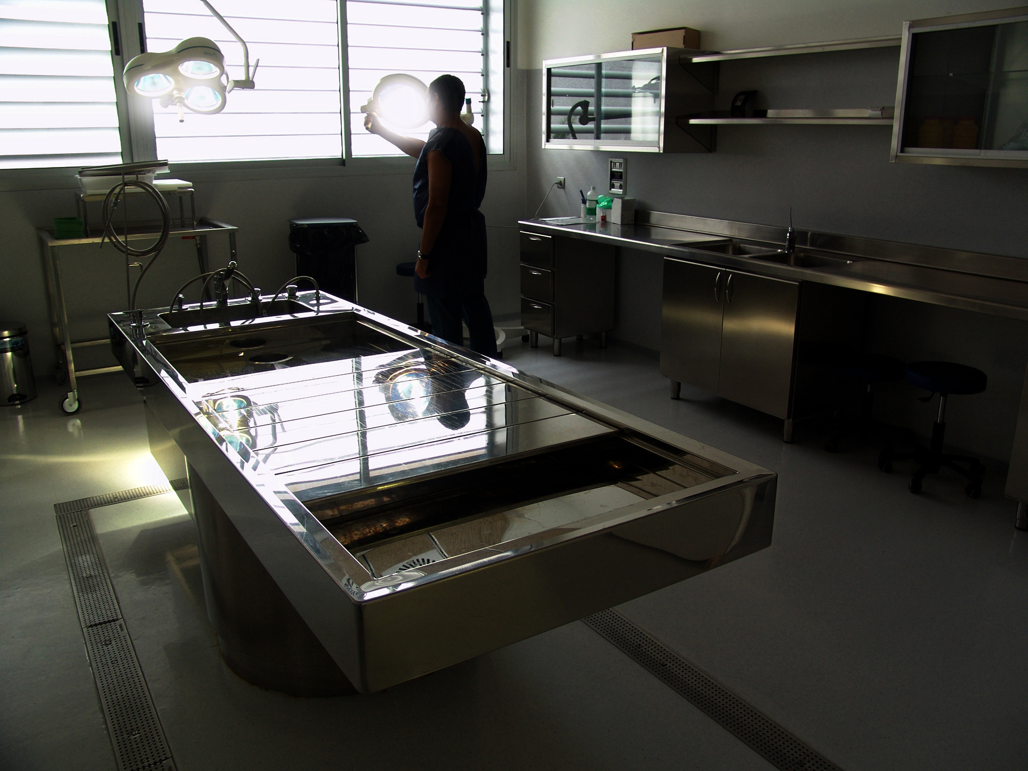 A person stands in a modern forensic laboratory with stainless steel equipment, including an autopsy table, under bright lights
