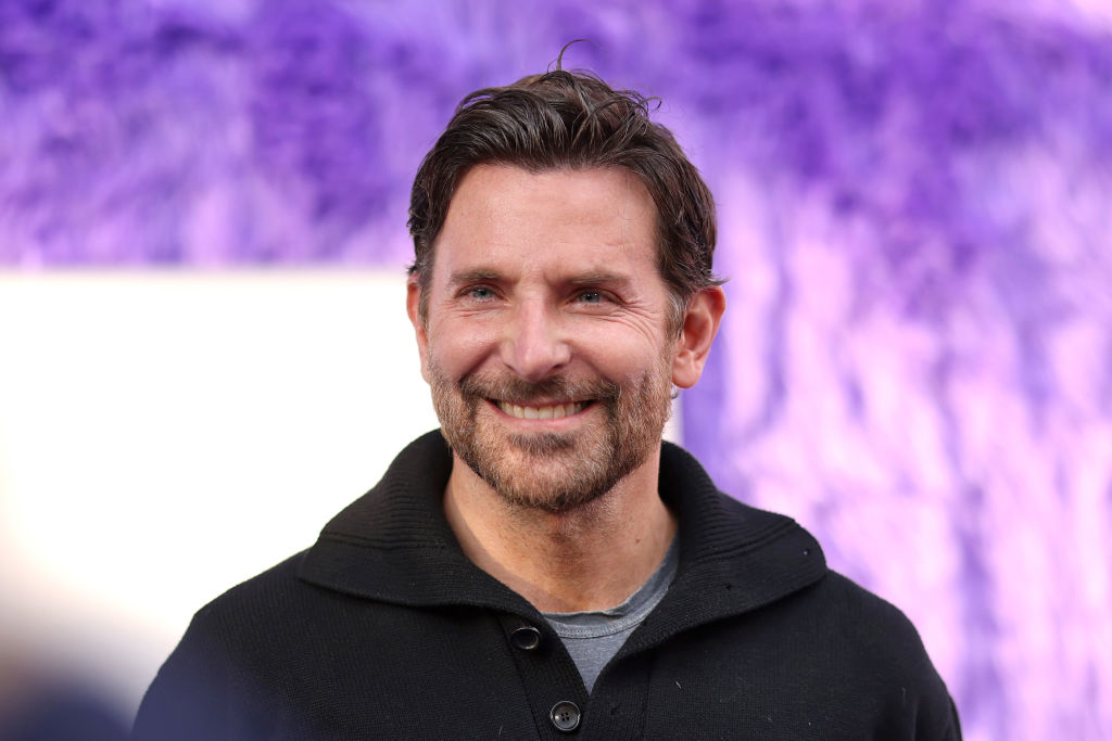 Bradley Cooper smiling at a public event, wearing a casual dark sweater with a gray shirt underneath