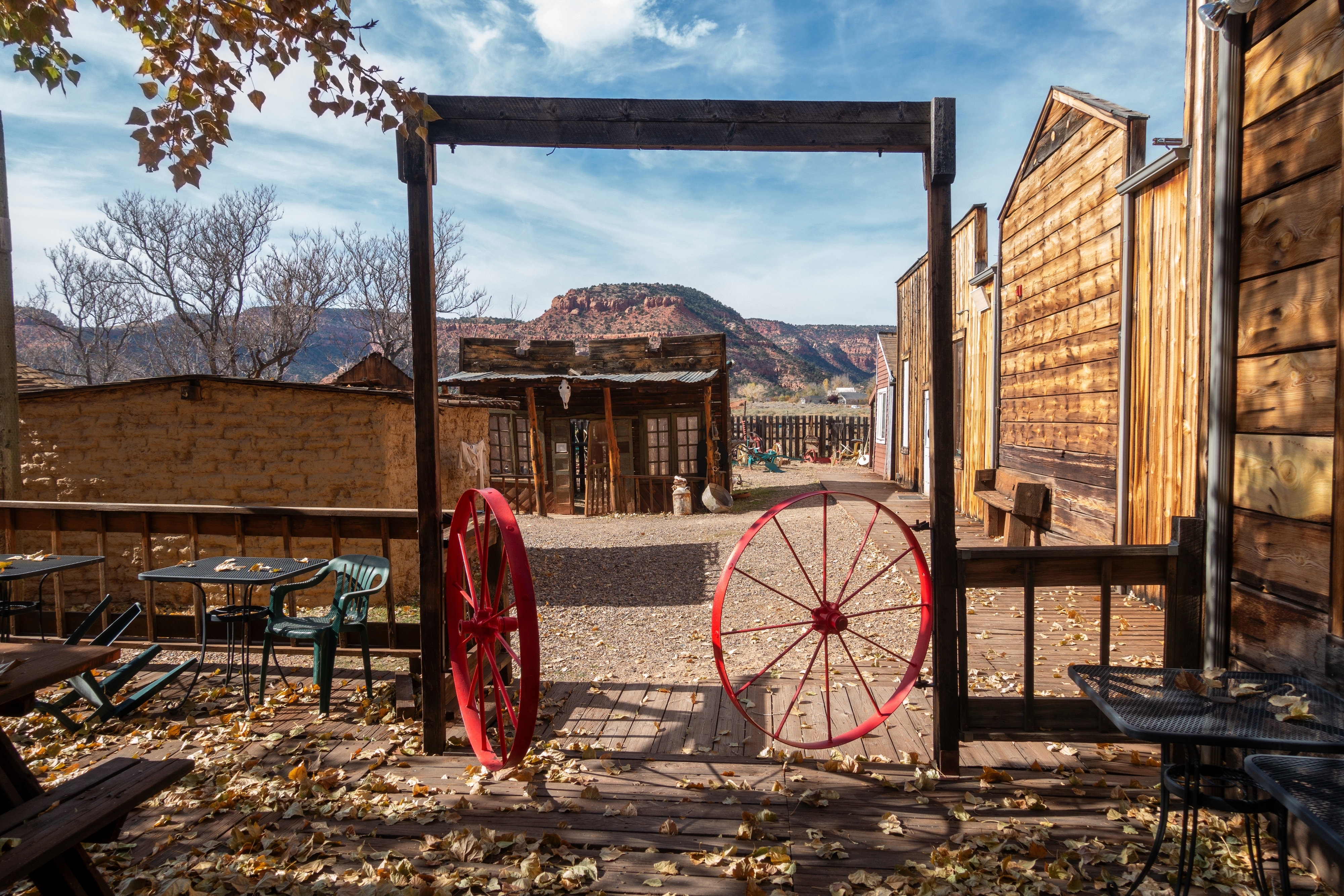 An old Western town scene with wooden buildings, two red wagon wheels at the entrance, some chairs and tables, with a mountainous landscape in the background