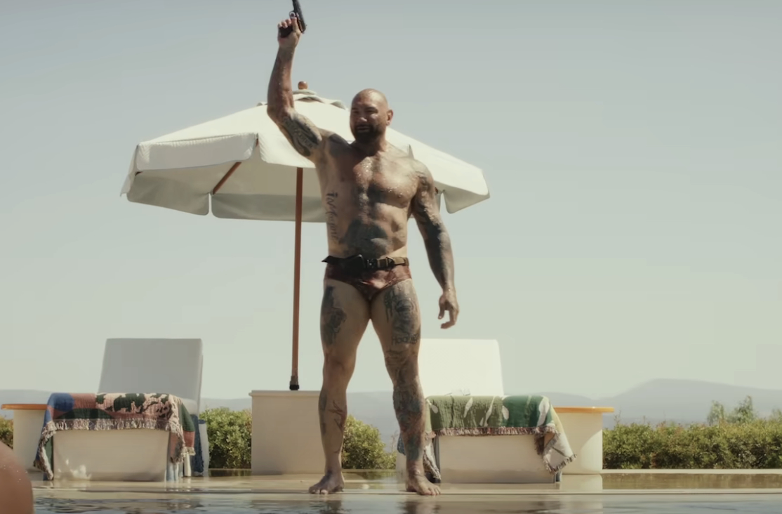 Dave Bautista stands shirtless and tattooed by a pool, holding a gun up, with an umbrella and pool chairs in the background